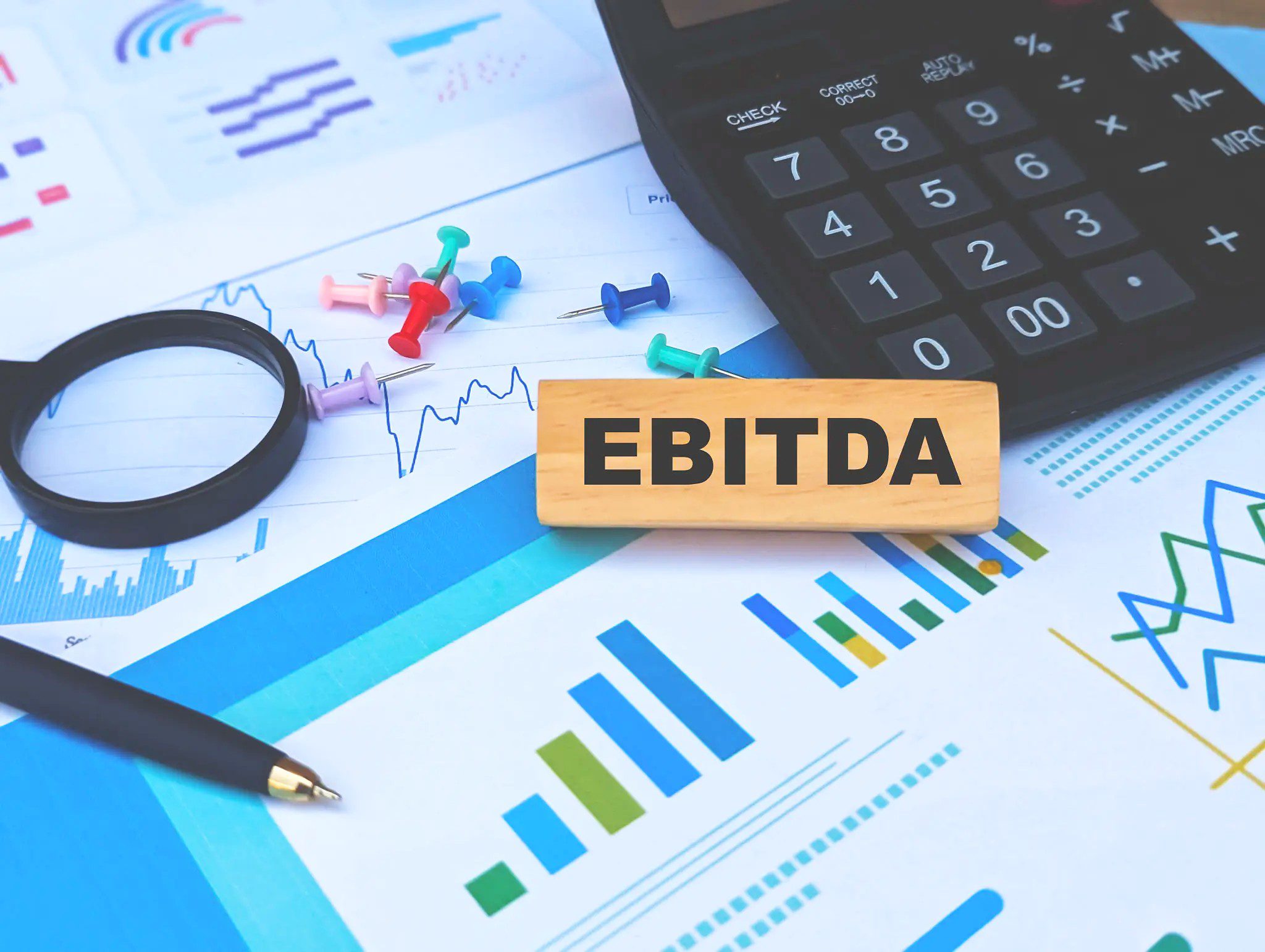 A wooden block labeled “EBITDA” sits on financial charts with a magnifying glass, pen, push pins, and calculator nearby.